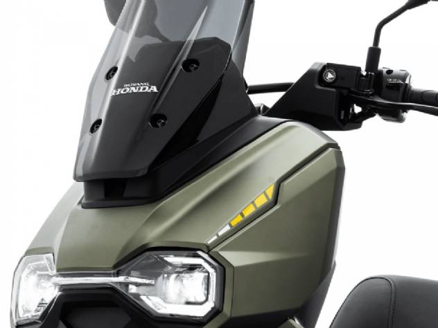 Honda HooRide 125 na cor cinza-chumbo, estacionada em um ambiente urbano com edifícios modernos ao fundo, exibindo seu design robusto, farol de LED e pneus de uso misto, pronta para o tráfego da cidade e pequenas trilhas. A imagem destaca a agilidade e o apelo aventureiro da scooter.