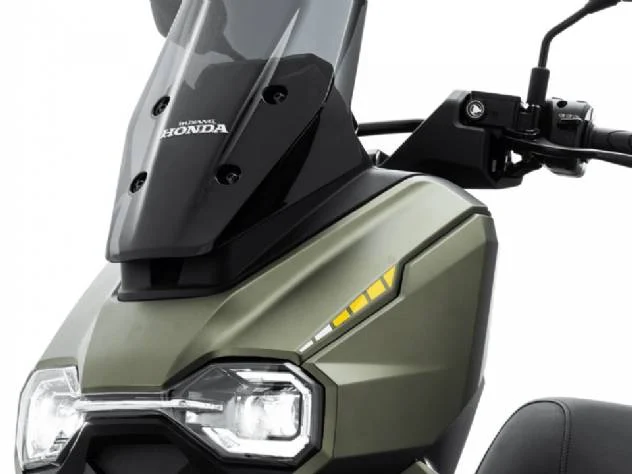 Honda HooRide 125 na cor cinza-chumbo, estacionada em um ambiente urbano com edifícios modernos ao fundo, exibindo seu design robusto, farol de LED e pneus de uso misto, pronta para o tráfego da cidade e pequenas trilhas. A imagem destaca a agilidade e o apelo aventureiro da scooter.