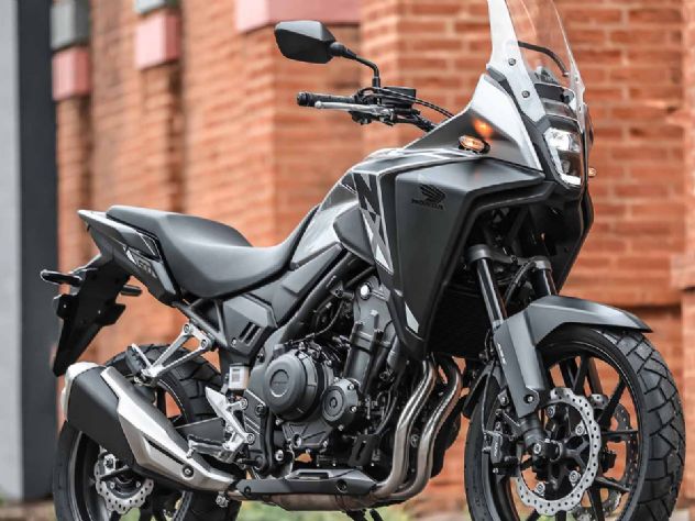 Uma motocicleta Honda NX500 2027 de cor vibrante, estacionada em uma estrada de terra com montanhas ao fundo, destacando seu design robusto e pneus de uso misto, pronta para aventura.