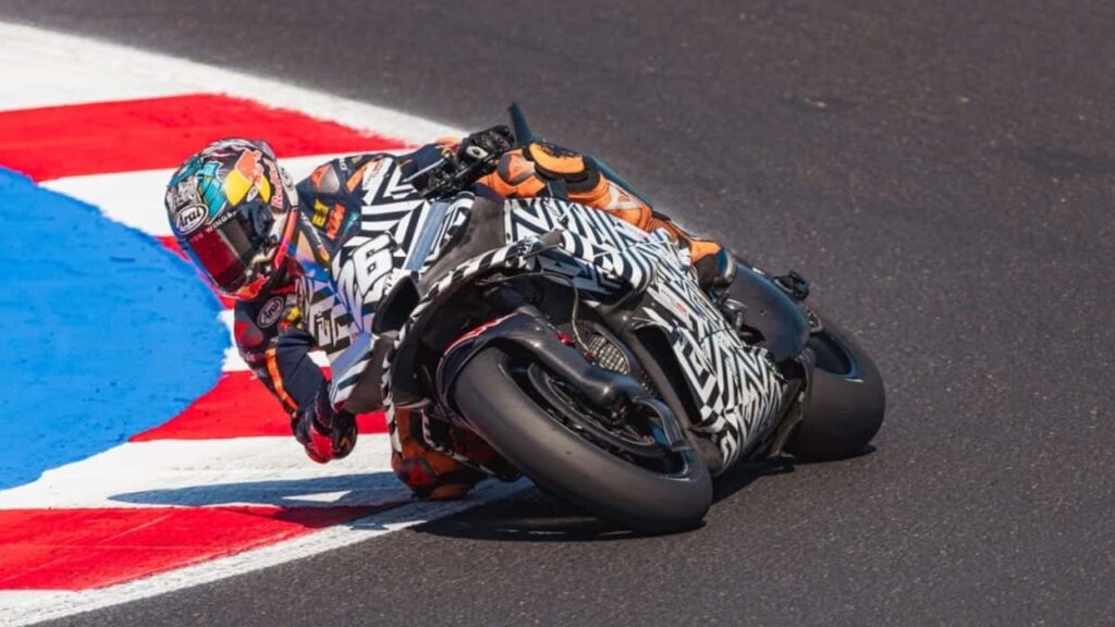 Protótipo da motocicleta KTM RC16 da próxima geração em testes na pista, destacando os novos elementos aerodinâmicos na carenagem, inspirados em designs de F1 e Aprilia, sob um céu ensolarado em um circuito de corrida.