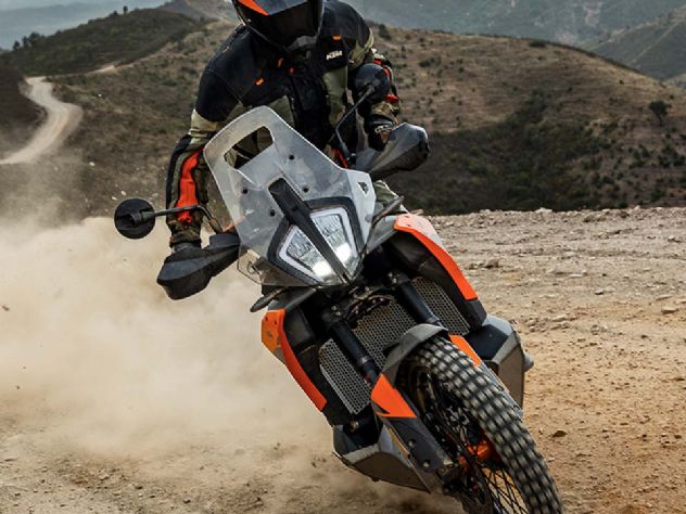 Vista lateral da motocicleta KTM 790 Adventure 2023 laranja e preta, estacionada em uma estrada de terra, com foco em seu design robusto e pneus de uso misto, pronta para aventura.