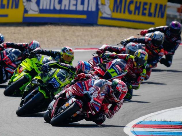 Motocicletas de MotoGP em alta velocidade durante uma corrida no icônico Circuito TT Assen, na Holanda, com pilotos se inclinando nas curvas.