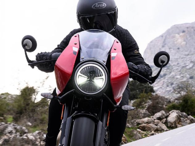 Uma Triumph Tracker 400 vermelha estacionada em uma paisagem urbana, com a silhueta de uma Thruxton 400 ao fundo, destacando a nova linha 400cc que chega ao Brasil.