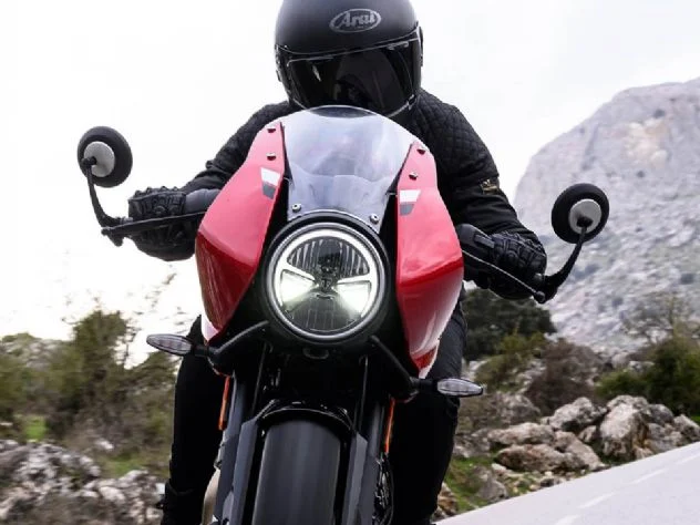 Uma Triumph Tracker 400 vermelha estacionada em uma paisagem urbana, com a silhueta de uma Thruxton 400 ao fundo, destacando a nova linha 400cc que chega ao Brasil.