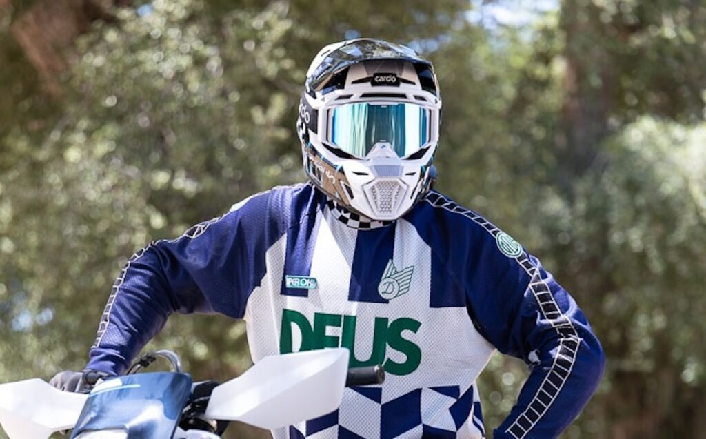 Capacete Cardo Venture na cor preta e vermelha, exibindo o sistema de comunicação integrado na lateral, em um cenário de trilha off-road com poeira ao fundo.