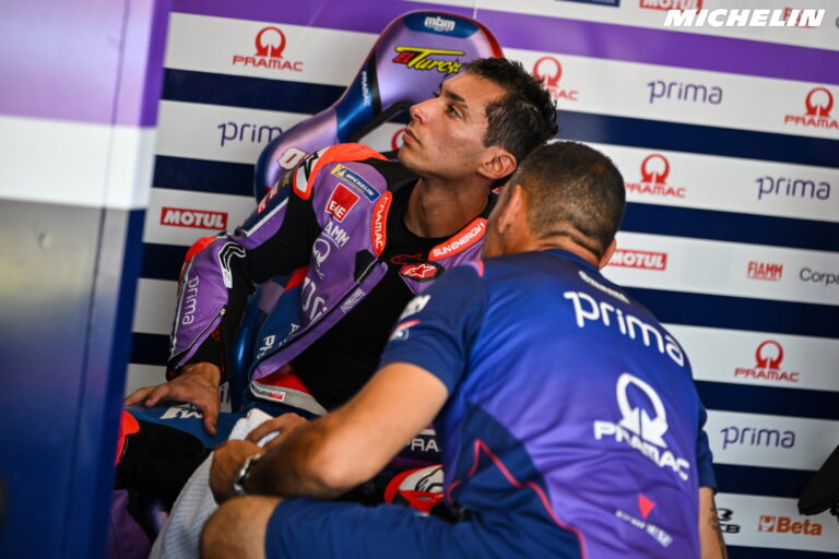 Toprak Razgatlioglu, piloto turco de motociclismo, inclinado em uma curva acentuada com sua motocicleta de corrida, com o joelho no chão, em alta velocidade durante uma competição. Ele usa um macacão de couro azul e branco, e o fundo mostra uma pista de corrida e arquibancadas.