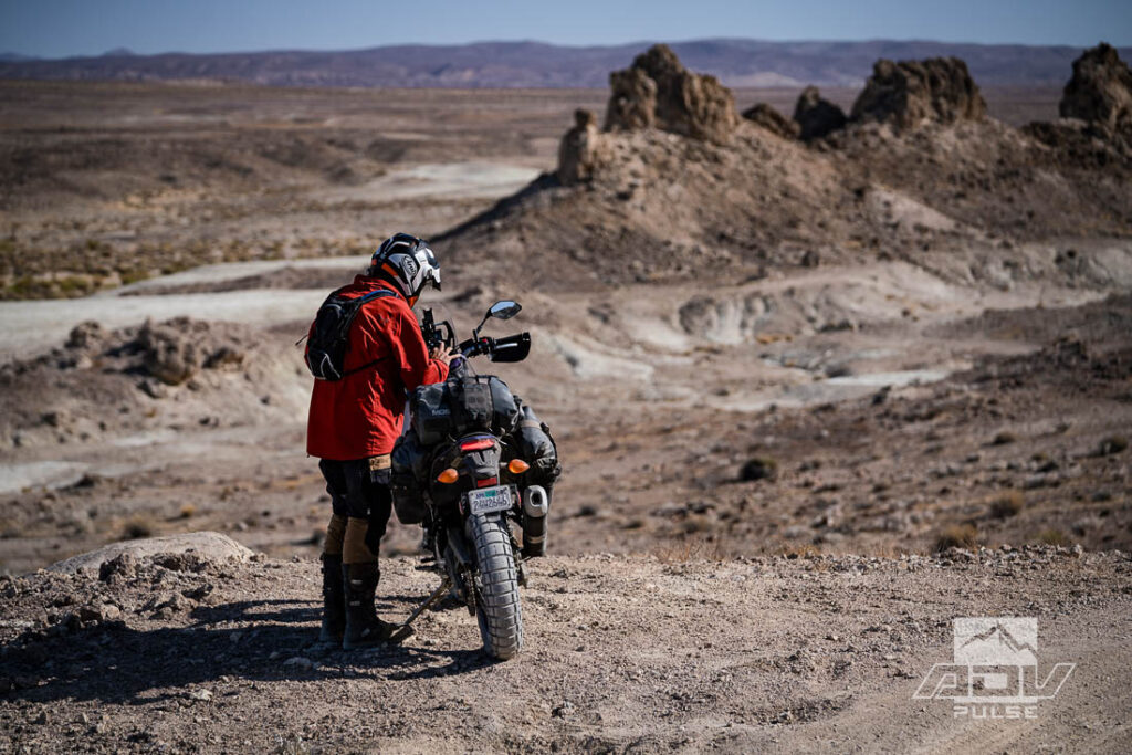 Motocicleta bigtrail estacionada entre as imponentes formações rochosas de tufa dos Trona Pinnacles, sob um céu azul e ensolarado no deserto de Mojave.