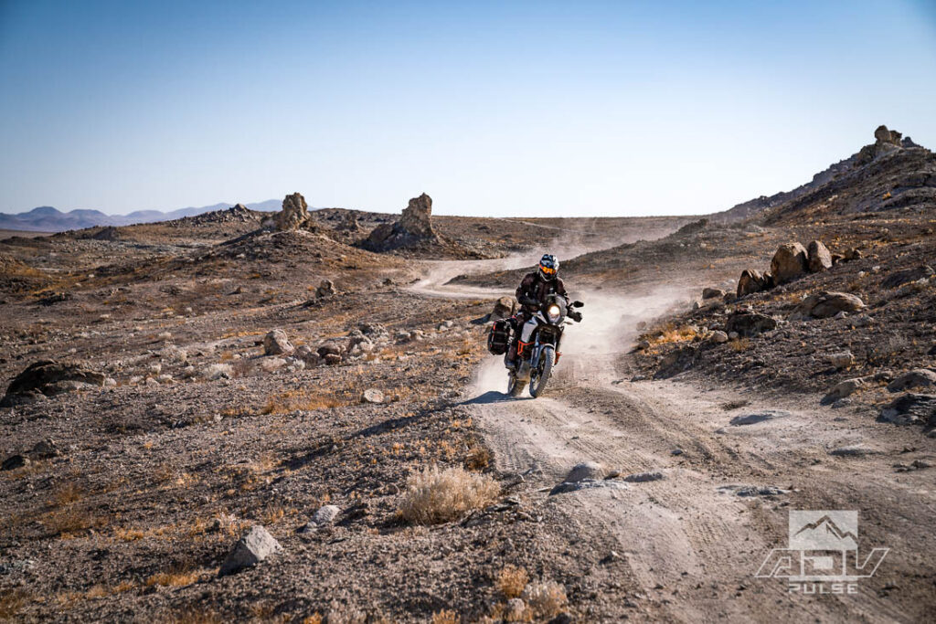 Motocicleta bigtrail estacionada entre as imponentes formações rochosas de tufa dos Trona Pinnacles, sob um céu azul e ensolarado no deserto de Mojave.