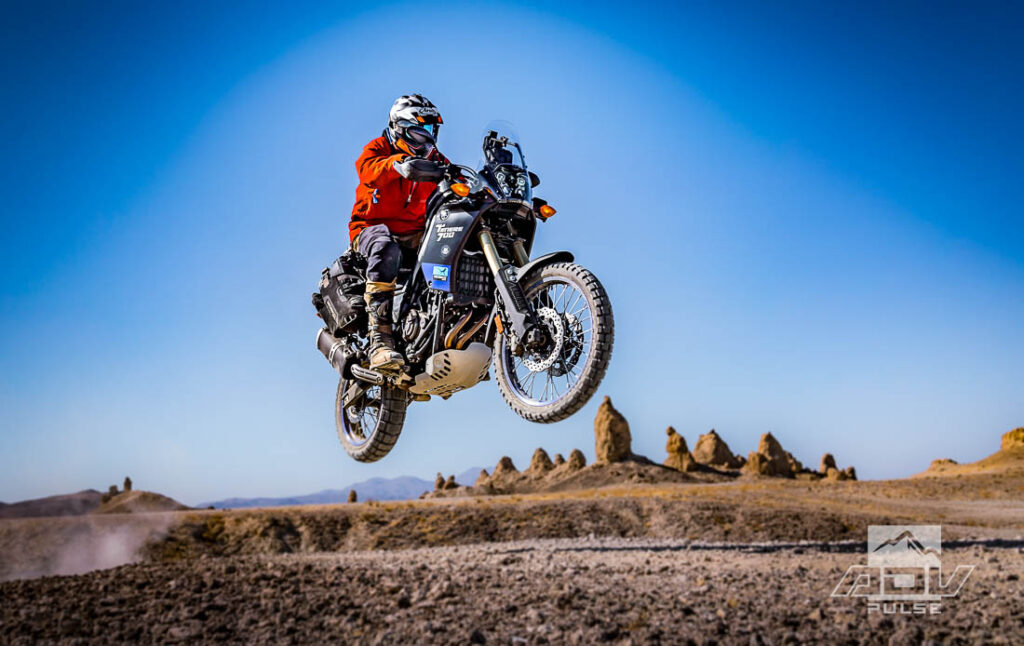 Motocicleta bigtrail estacionada entre as imponentes formações rochosas de tufa dos Trona Pinnacles, sob um céu azul e ensolarado no deserto de Mojave.