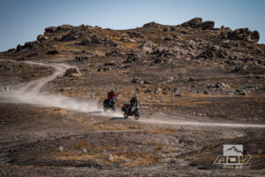 Motocicleta bigtrail estacionada entre as imponentes formações rochosas de tufa dos Trona Pinnacles, sob um céu azul e ensolarado no deserto de Mojave.