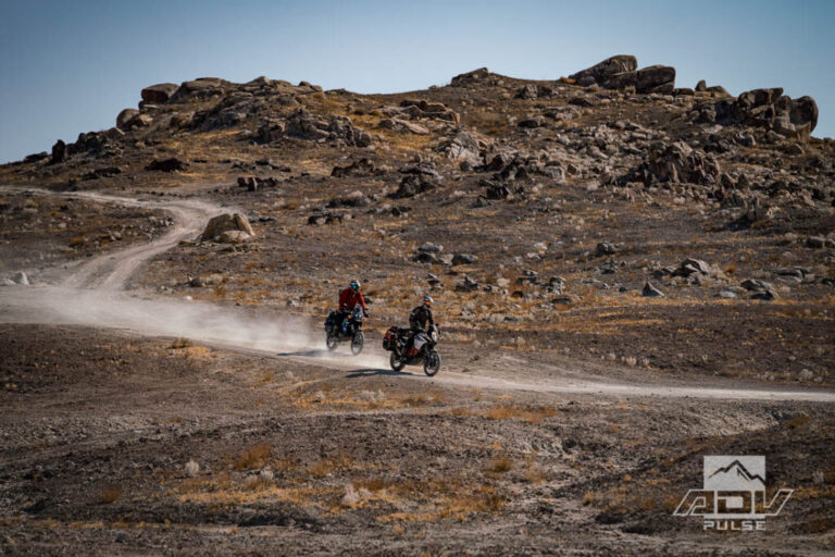 Motocicleta bigtrail estacionada entre as imponentes formações rochosas de tufa dos Trona Pinnacles, sob um céu azul e ensolarado no deserto de Mojave.
