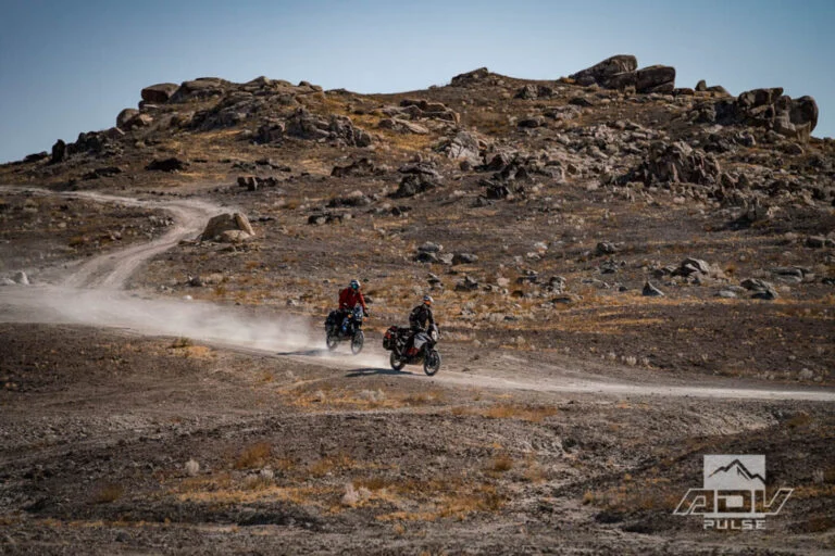 Motocicleta bigtrail estacionada entre as imponentes formações rochosas de tufa dos Trona Pinnacles, sob um céu azul e ensolarado no deserto de Mojave.
