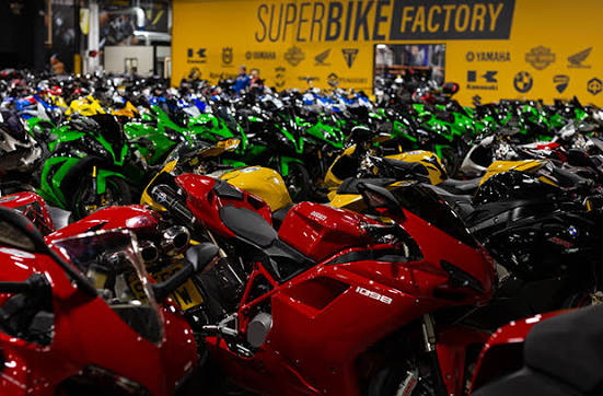 Vista panorâmica de uma grande concessionária de motocicletas usadas, SuperBike Factory, com centenas de motos de diversas marcas e modelos expostas no showroom. Detalhe da organização e iluminação do espaço.