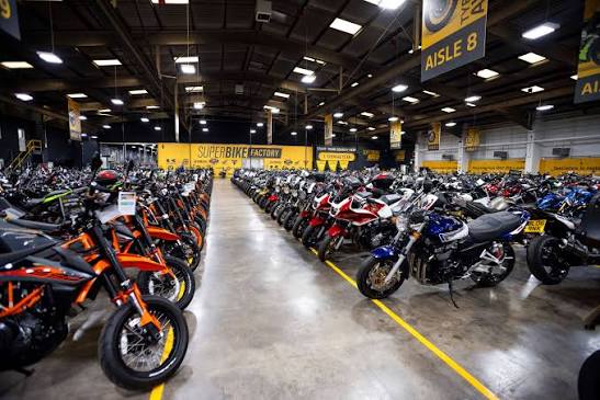 Vista panorâmica de uma grande concessionária de motocicletas usadas, SuperBike Factory, com centenas de motos de diversas marcas e modelos expostas no showroom. Detalhe da organização e iluminação do espaço.