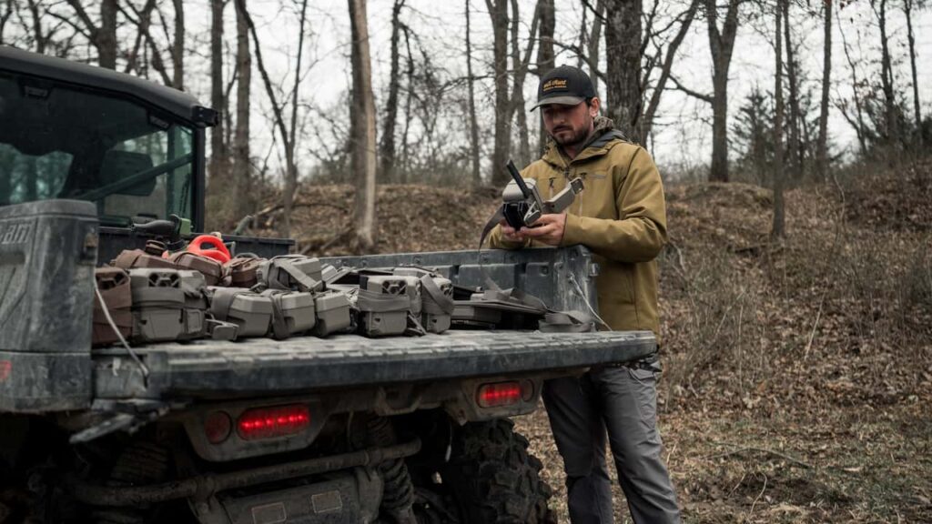 Chris Bee, um arqueiro e caçador profissional, posa ao lado de um UTV elétrico Can-Am Maverick em um cenário de floresta, destacando a capacidade off-road e a tecnologia sustentável do veículo.
