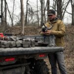 Chris Bee, um arqueiro e caçador profissional, posa ao lado de um UTV elétrico Can-Am Maverick em um cenário de floresta, destacando a capacidade off-road e a tecnologia sustentável do veículo.