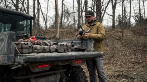 Chris Bee, um arqueiro e caçador profissional, posa ao lado de um UTV elétrico Can-Am Maverick em um cenário de floresta, destacando a capacidade off-road e a tecnologia sustentável do veículo.