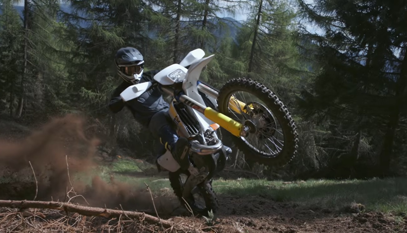 Piloto de enduro em ação com uma moto Husqvarna Enduro 2027 vermelha e branca, enfrentando um trilho técnico rochoso em alta velocidade, demonstrando a precisão do novo modelo.