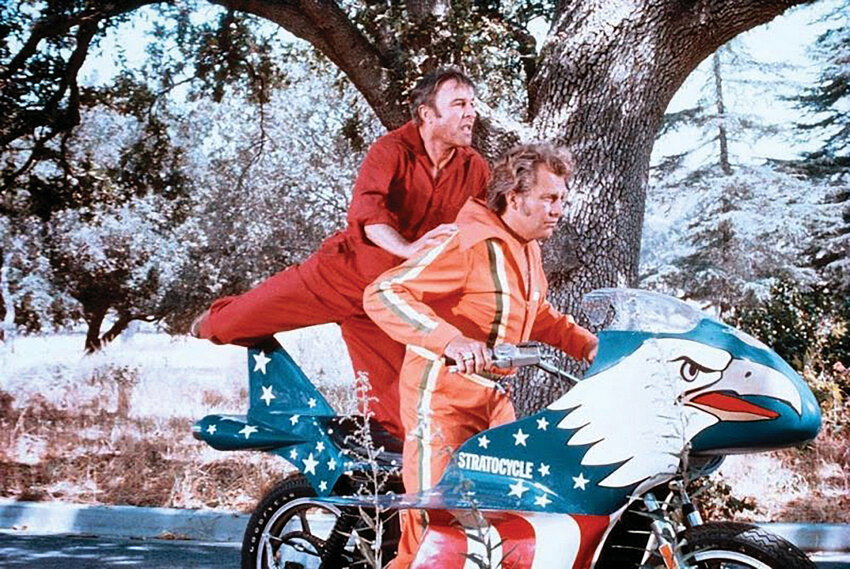 Minibike Ruttman 1972 restaurada, vermelha e branca, em exibição, com estilo remetendo aos anos 70 e ao legado de Evel Knievel.