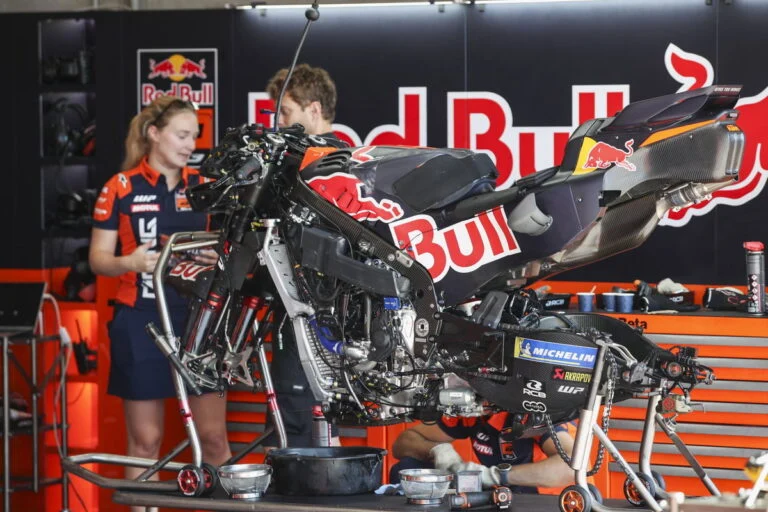 Duas motos KTM RC16 da equipe Red Bull KTM Factory Racing em plena ação na pista de MotoGP, mostrando a potência e a aerodinâmica das máquinas de corrida, mas enfrentando problemas de motor.