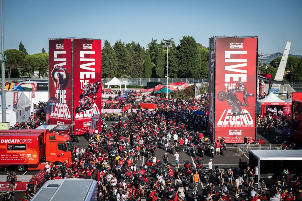 Vista aérea do paddock do Circuito de Misano, com milhares de motocicletas Ducati estacionadas e entusiastas caminhando durante a World Ducati Week, sob um céu azul.