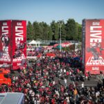 Vista aérea do paddock do Circuito de Misano, com milhares de motocicletas Ducati estacionadas e entusiastas caminhando durante a World Ducati Week, sob um céu azul.
