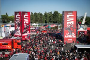 Vista aérea do paddock do Circuito de Misano, com milhares de motocicletas Ducati estacionadas e entusiastas caminhando durante a World Ducati Week, sob um céu azul.