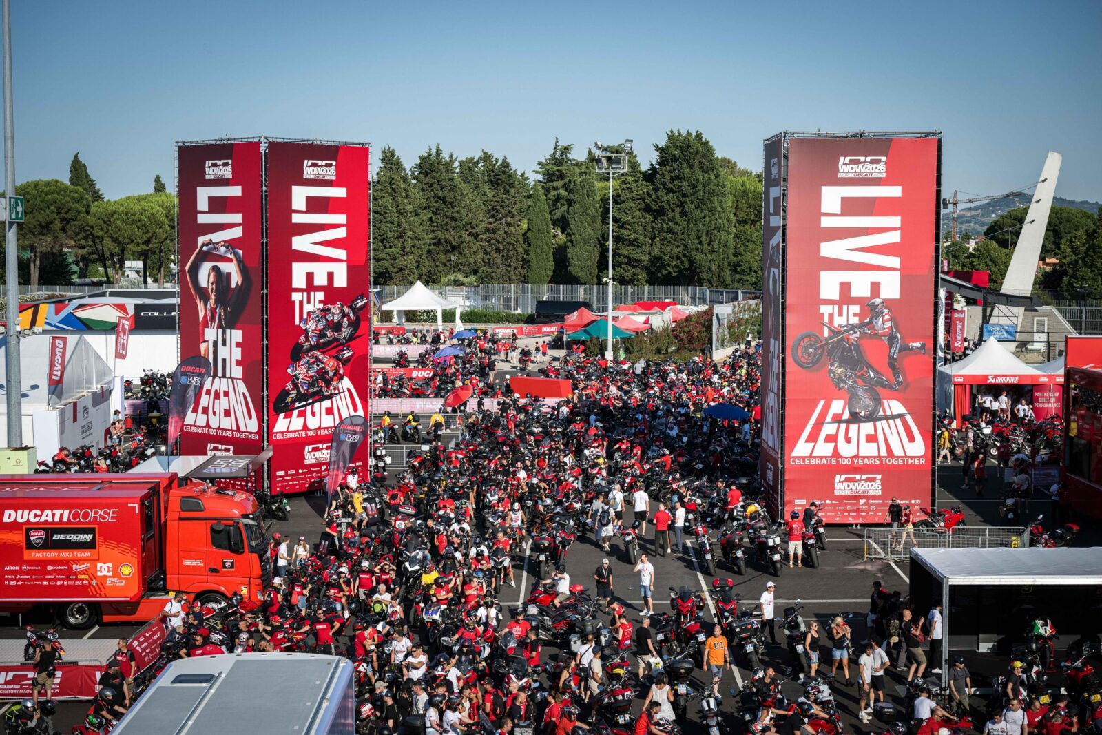 Vista aérea do paddock do Circuito de Misano, com milhares de motocicletas Ducati estacionadas e entusiastas caminhando durante a World Ducati Week, sob um céu azul.