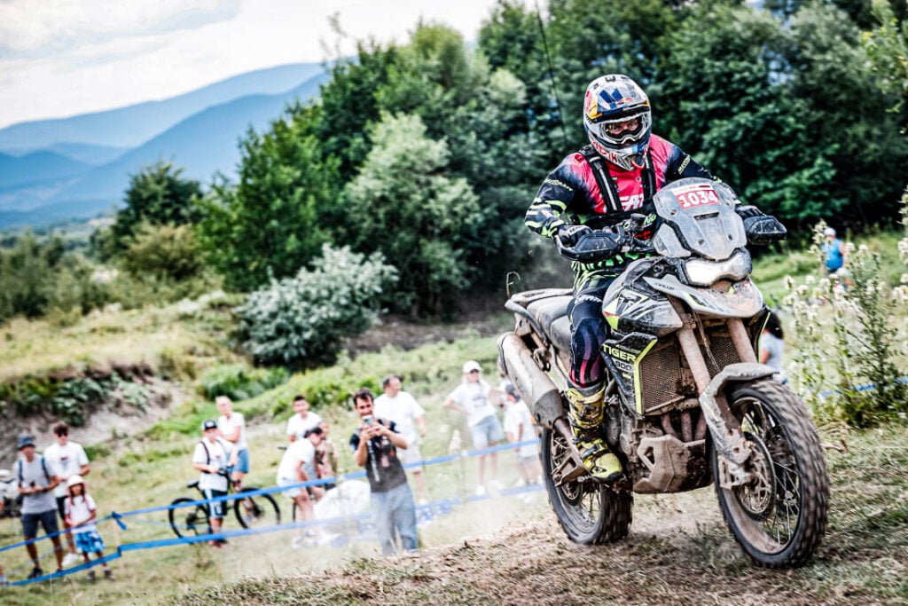 Uma Triumph Tiger 900 Rally Pro em ação em uma trilha técnica de hard enduro, com um piloto experiente. Foco na robustez da motocicleta.