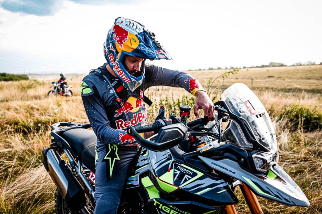 Uma Triumph Tiger 900 Rally Pro em ação em uma trilha técnica de hard enduro, com um piloto experiente. Foco na robustez da motocicleta.
