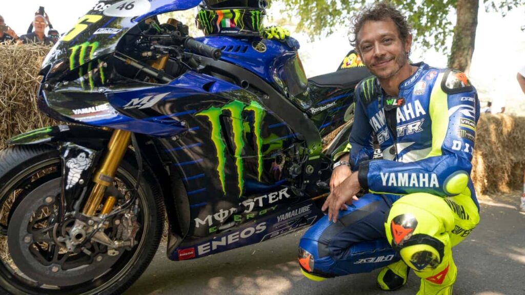 Valentino Rossi pilotando sua icônica Yamaha M1 azul e branca em alta velocidade na pista do Festival de Goodwood, com fãs ao fundo.