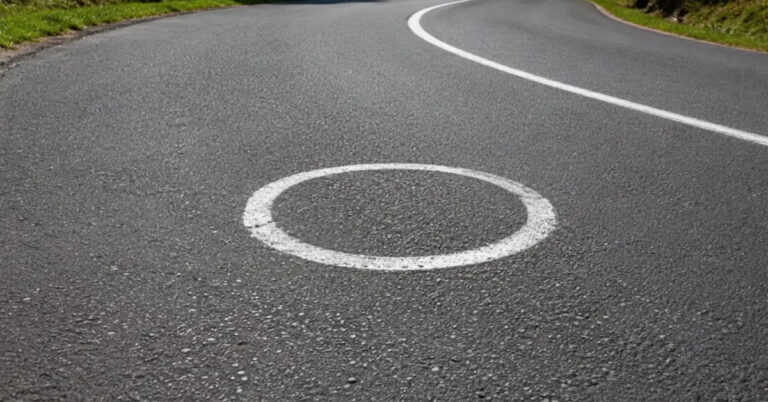 Círculos brancos pintados no asfalto de uma estrada sinuosa na Espanha, visando melhorar a trajetória e a segurança de motociclistas em curvas perigosas.