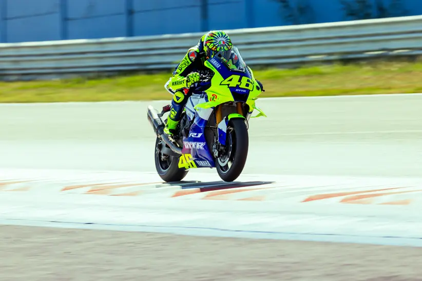 Valentino Rossi e outros pilotos de MotoGP ao lado de engenheiros da Dainese, analisando um macacão de segurança no circuito de Misano durante o Project Apex 2026.