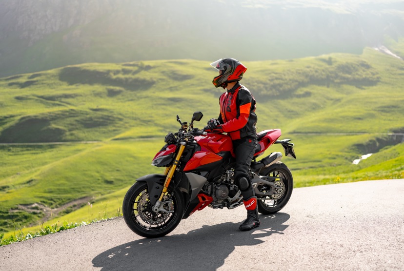 Motociclista vestindo equipamento completo Ducati, incluindo calças e botas, em uma moto esportiva na estrada.