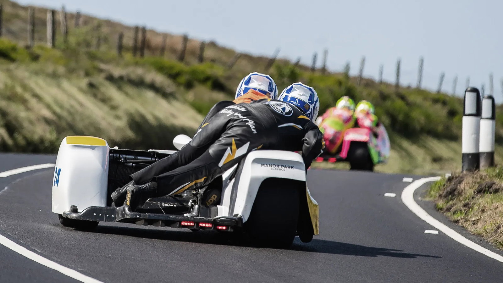 Foto de um sidecar em alta velocidade na Isle of Man TT, com piloto e passageiro em perfeita sincronia, contornando uma curva fechada no famoso circuito da ilha. O cenário de casas e árvores passa em um borrão, destacando a velocidade e a emoção da corrida.