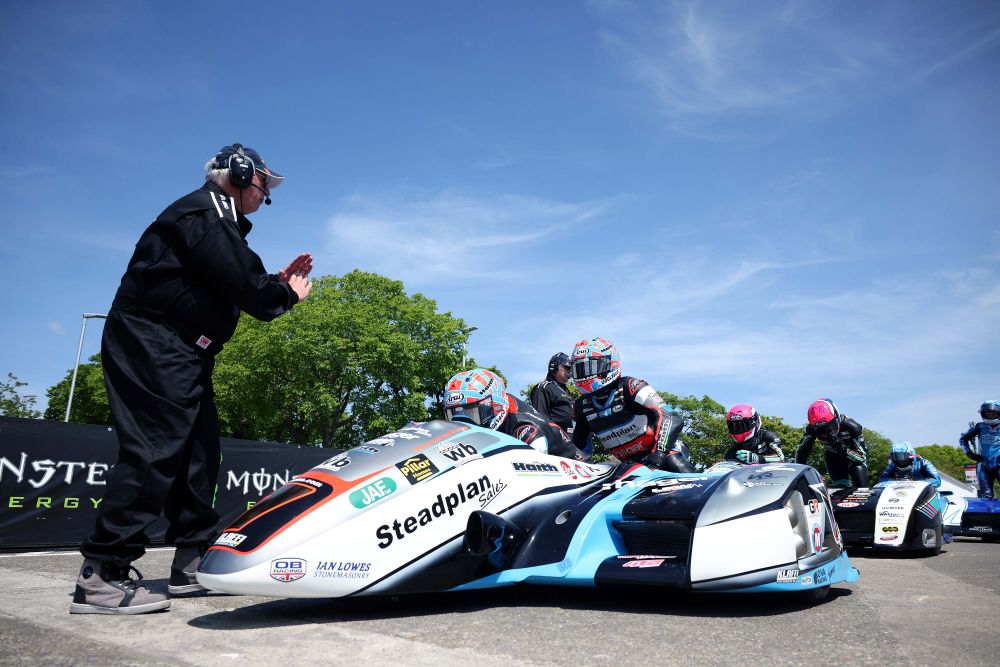 Foto de um sidecar em alta velocidade na Isle of Man TT, com piloto e passageiro em perfeita sincronia, contornando uma curva fechada no famoso circuito da ilha. O cenário de casas e árvores passa em um borrão, destacando a velocidade e a emoção da corrida.