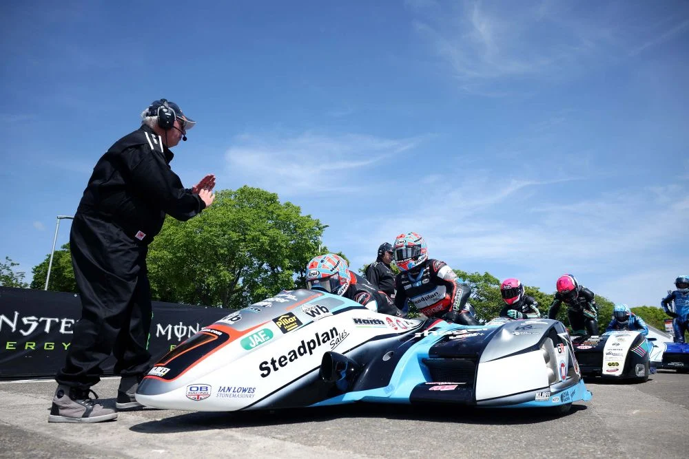 Foto de um sidecar em alta velocidade na Isle of Man TT, com piloto e passageiro em perfeita sincronia, contornando uma curva fechada no famoso circuito da ilha. O cenário de casas e árvores passa em um borrão, destacando a velocidade e a emoção da corrida.
