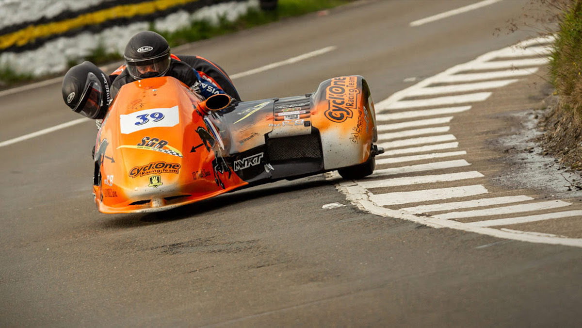 Foto de um sidecar em alta velocidade na Isle of Man TT, com piloto e passageiro em perfeita sincronia, contornando uma curva fechada no famoso circuito da ilha. O cenário de casas e árvores passa em um borrão, destacando a velocidade e a emoção da corrida.