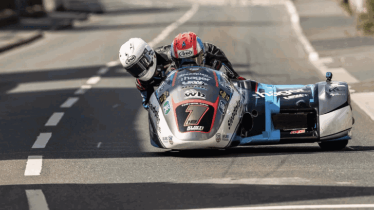 Foto de um sidecar em alta velocidade na Isle of Man TT, com piloto e passageiro em perfeita sincronia, contornando uma curva fechada no famoso circuito da ilha. O cenário de casas e árvores passa em um borrão, destacando a velocidade e a emoção da corrida.