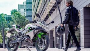 Uma Kawasaki Ninja e-1 na cor verde limão e preta, com design esportivo característico da linha Ninja, estacionada em um ambiente urbano. Destaca a carenagem aerodinâmica e os detalhes elétricos do modelo de 2027.