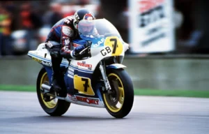 Barry Sheene pilotando a icônica Suzuki RG500 de dois tempos durante uma corrida de Grande Prêmio na década de 1970, destacando a agressividade e velocidade da máquina.