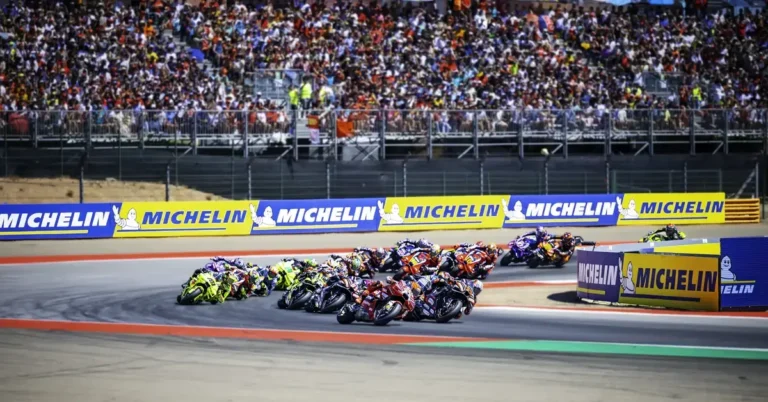 Piloto de MotoGP em alta velocidade na pista, com sistema de rádio visível no capacete, ilustrando a futura comunicação entre equipes e pilotos.