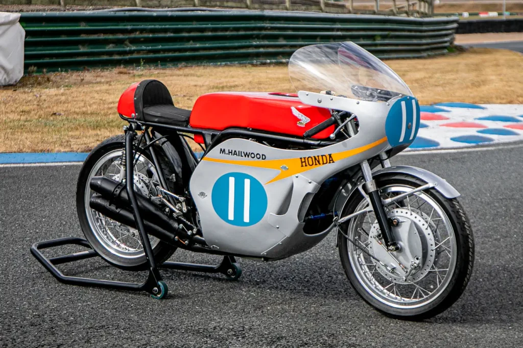 A lendária Honda RC173, uma motocicleta de corrida de fábrica de 350cc com quatro cilindros, pilotada por Mike Hailwood, exibida em um evento. A moto de cor vermelha e branca está estacionada, destacando seu design clássico de corrida dos anos 60 e o número 1 estampado.