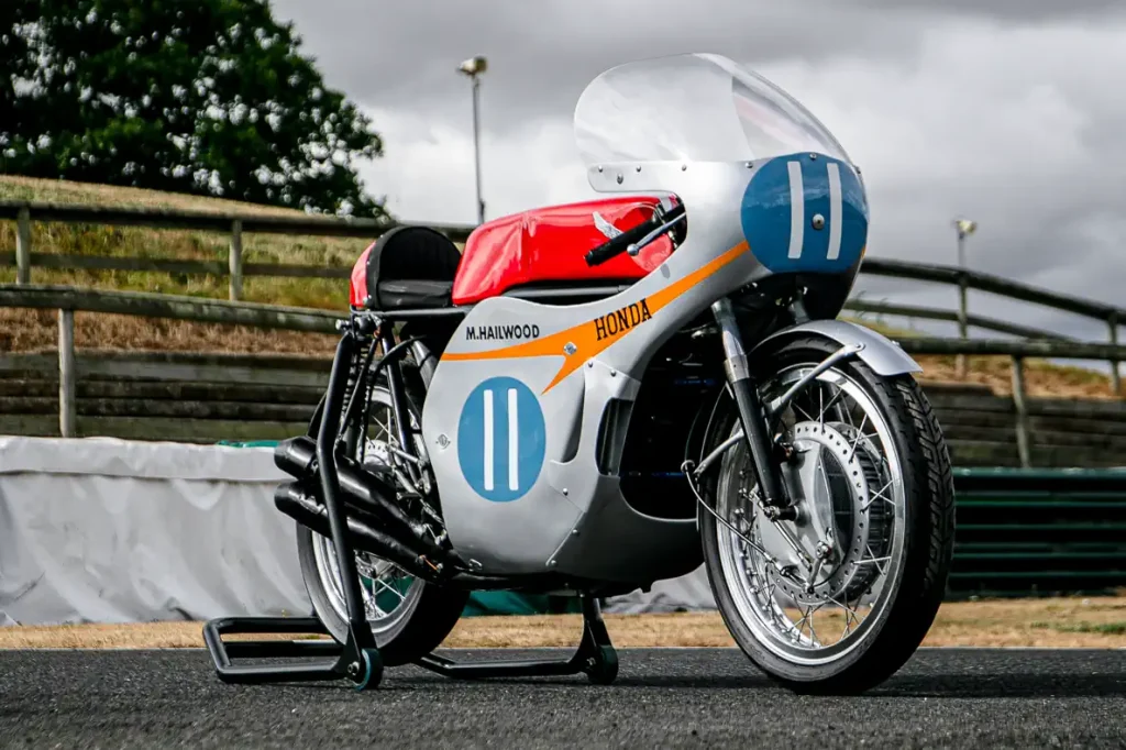 A lendária Honda RC173, uma motocicleta de corrida de fábrica de 350cc com quatro cilindros, pilotada por Mike Hailwood, exibida em um evento. A moto de cor vermelha e branca está estacionada, destacando seu design clássico de corrida dos anos 60 e o número 1 estampado.