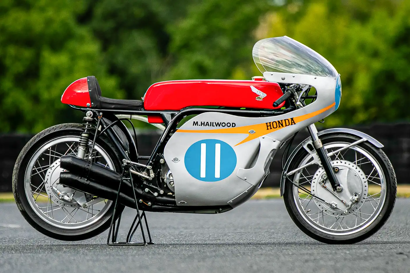 A lendária Honda RC173, uma motocicleta de corrida de fábrica de 350cc com quatro cilindros, pilotada por Mike Hailwood, exibida em um evento. A moto de cor vermelha e branca está estacionada, destacando seu design clássico de corrida dos anos 60 e o número 1 estampado.