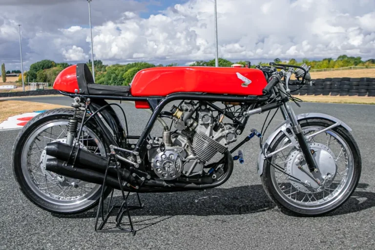 A lendária Honda RC173, uma motocicleta de corrida de fábrica de 350cc com quatro cilindros, pilotada por Mike Hailwood, exibida em um evento. A moto de cor vermelha e branca está estacionada, destacando seu design clássico de corrida dos anos 60 e o número 1 estampado.