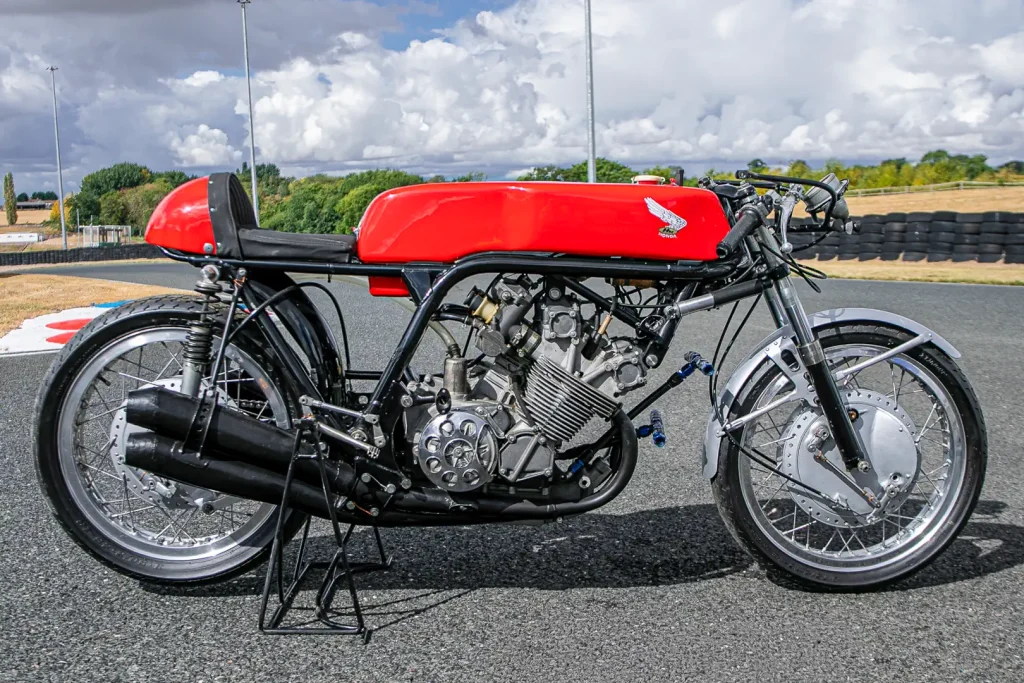 A lendária Honda RC173, uma motocicleta de corrida de fábrica de 350cc com quatro cilindros, pilotada por Mike Hailwood, exibida em um evento. A moto de cor vermelha e branca está estacionada, destacando seu design clássico de corrida dos anos 60 e o número 1 estampado.