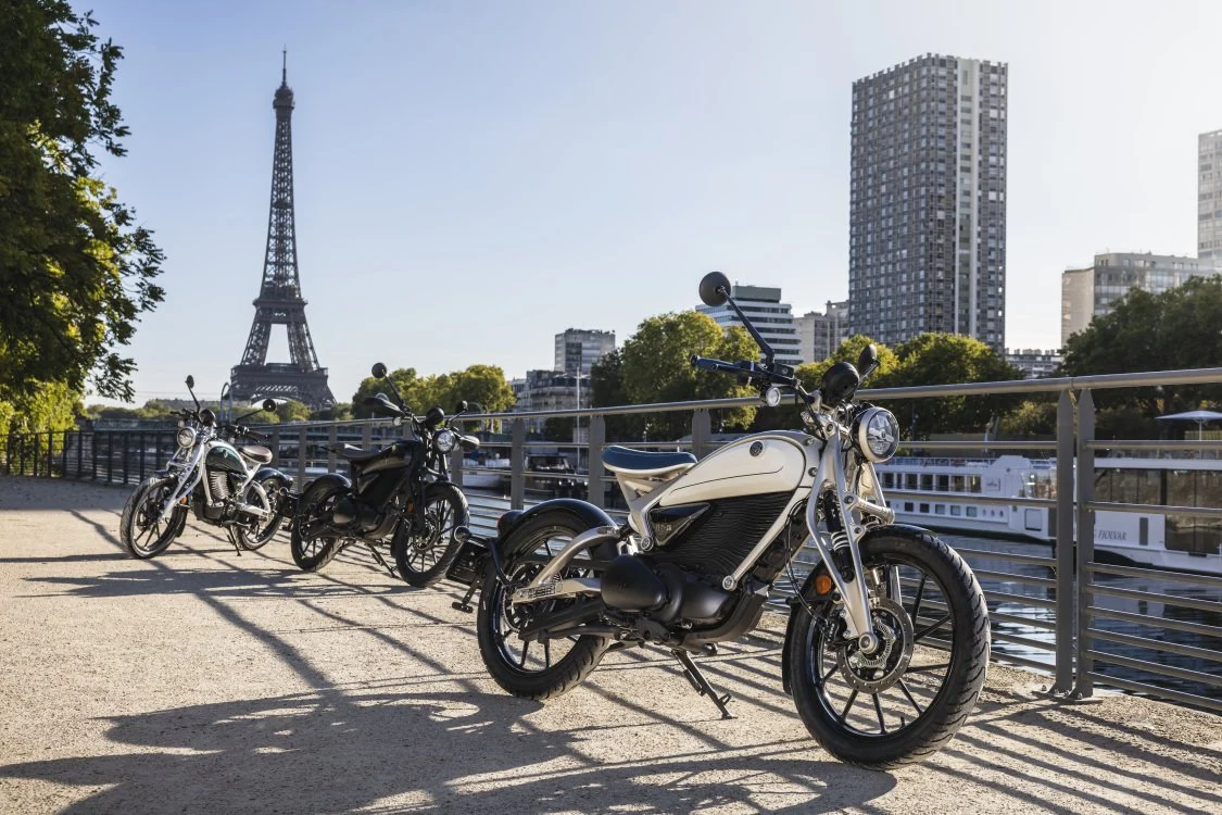 Motocicleta elétrica Royal Enfield Flying Flea C6 em destaque, exibindo seu design moderno e compacto, com farol redondo e suspensão dianteira girder, em ambiente urbano.