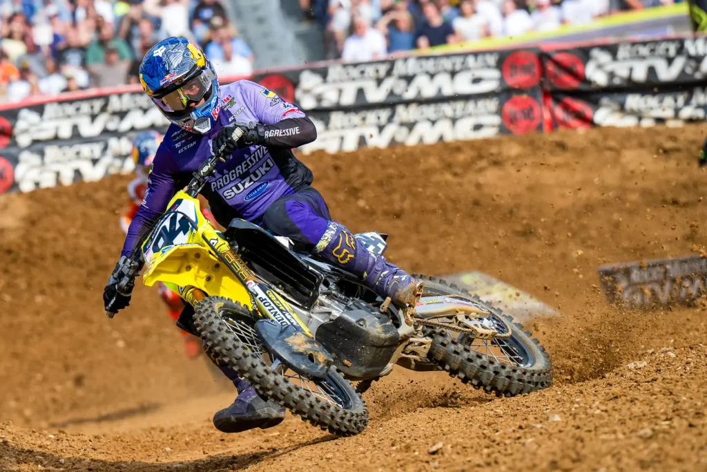 Detalhe de um piloto de SuperMotocross em ação, saltando sobre um obstáculo com a moto no ar, em um estádio lotado.