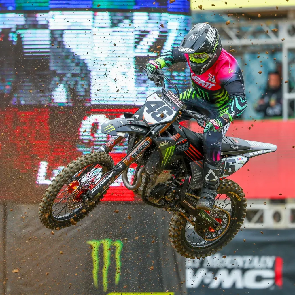 Detalhe de um piloto de SuperMotocross em ação, saltando sobre um obstáculo com a moto no ar, em um estádio lotado.